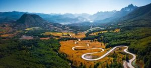 Aerial view of the winding Carretera Austral road in Chile, surrounded by lush green forests, golden meadows, and distant mountains under a clear blue sky.