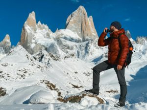 A hiker in a red jacket and black beanie stands confidently on a snowy rock, drinking water against the backdrop of the majestic Fitz Roy mountain in Patagonia, Argentina. The bright blue sky contrasts with the white snow-covered peaks.