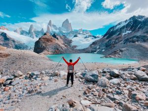 A hiker raising trekking poles in triumph while standing by the turquoise waters of Laguna de los Tres, with the majestic Mount Fitz Roy in the background under a vibrant blue sky. The rocky terrain and glacial surroundings highlight the breathtaking beauty of Patagonia, Argentina.