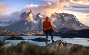 A hiker dressed in an orange jacket standing on a rocky outcrop, admiring the stunning Torres del Paine mountains in Chile. The scene features dramatic peaks illuminated by golden sunset light, with turquoise lakes and rugged terrain characteristic of Patagonia.