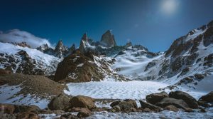 Breathtaking view of Mount Fitz Roy in Patagonia, with its rugged peaks piercing a clear blue sky. A snow-covered glacial landscape stretches across the foreground, bordered by rocky terrain, highlighting the dramatic and pristine beauty of the region under the midday sun