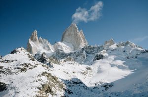 A stunning view of Mount Fitz Roy in Patagonia, Argentina, towering over a pristine snow-covered landscape under a bright blue sky. The jagged peaks and untouched snow highlight the majestic and rugged beauty of the Andes mountains.