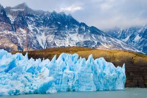 A breathtaking view of the Perito Moreno Glacier in Patagonia, Argentina, featuring vibrant blue jagged ice formations stretching across the foreground. Behind the glacier, towering snow-dusted mountains rise dramatically under a moody, overcast sky, emphasizing the region's wild and pristine beauty.