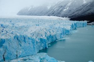 Close-up view of the Perito Moreno Glacier in Patagonia, showcasing its towering blue ice walls meeting a tranquil turquoise lake. The glacier's intricate patterns of ice formations contrast with the snow-dusted mountains and cloudy sky, creating a breathtaking natural scene.