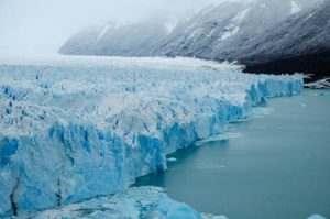 The majestic Perito Moreno Glacier in Los Glaciares National Park, Argentina. Its icy blue wall towers over the calm turquoise waters, with snow-covered mountains visible in the background under a misty sky.