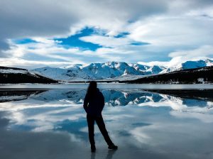 Silhouette of a person standing on the icy surface of a reflective lake in Patagonia, surrounded by snow-covered mountains under a dramatic cloudy sky. The tranquil lake mirrors the pristine peaks and sky, creating a stunning and serene winter landscape.