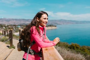 A woman in hiking gear enjoys a sunny coastal view, embodying outdoor adventure.