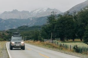 An SUV on a winding road in Puerto Montt, Chile, surrounded by lush green forests and majestic mountains partially covered in mist and snow, capturing the essence of a scenic road trip adventure.